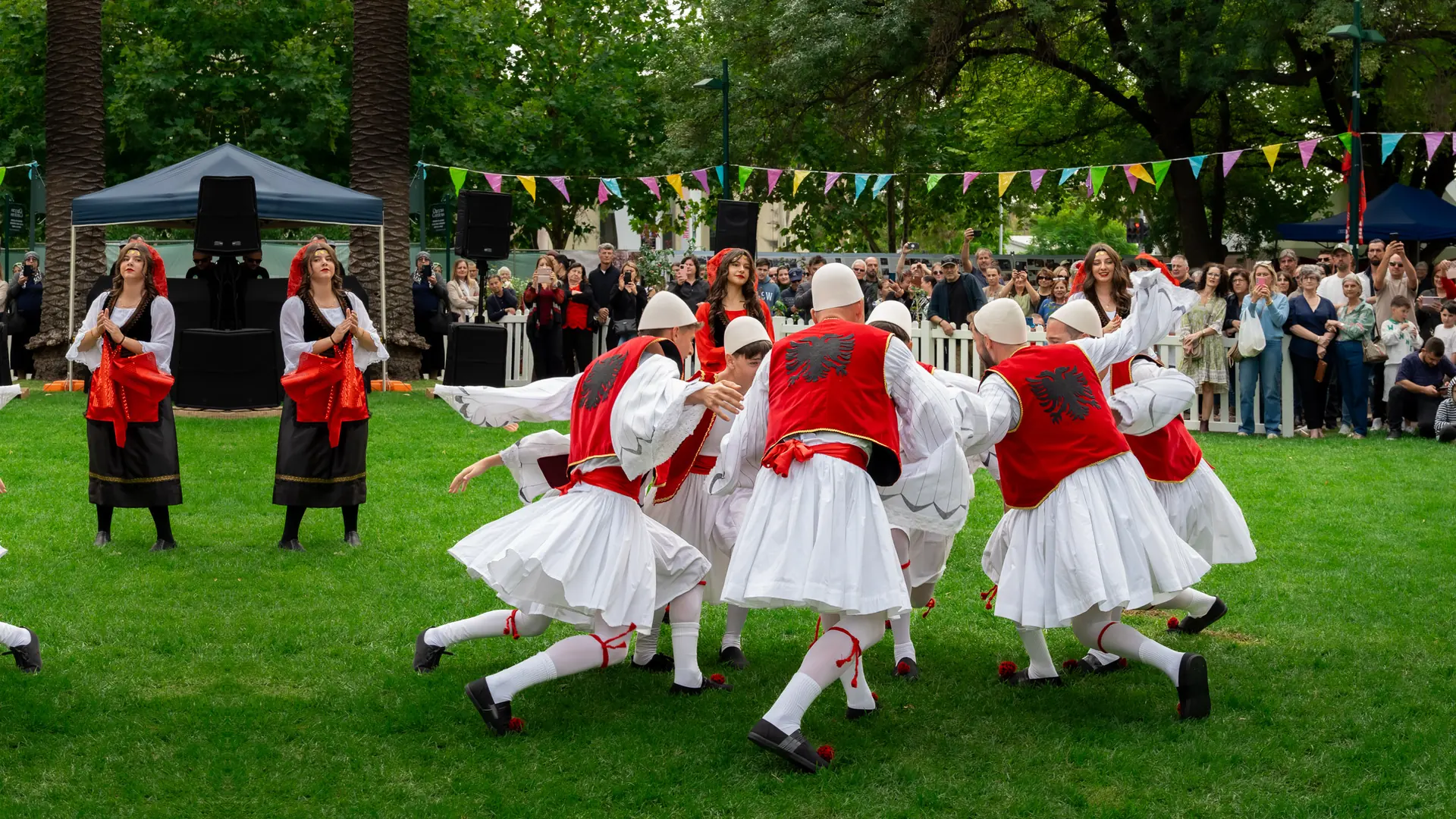Men in white pleated skirts, red waistcoats with a black double-headed eagle and white felt caps dance in a tight ring on grass while a crowd watches and women in red and black costume stand beside them