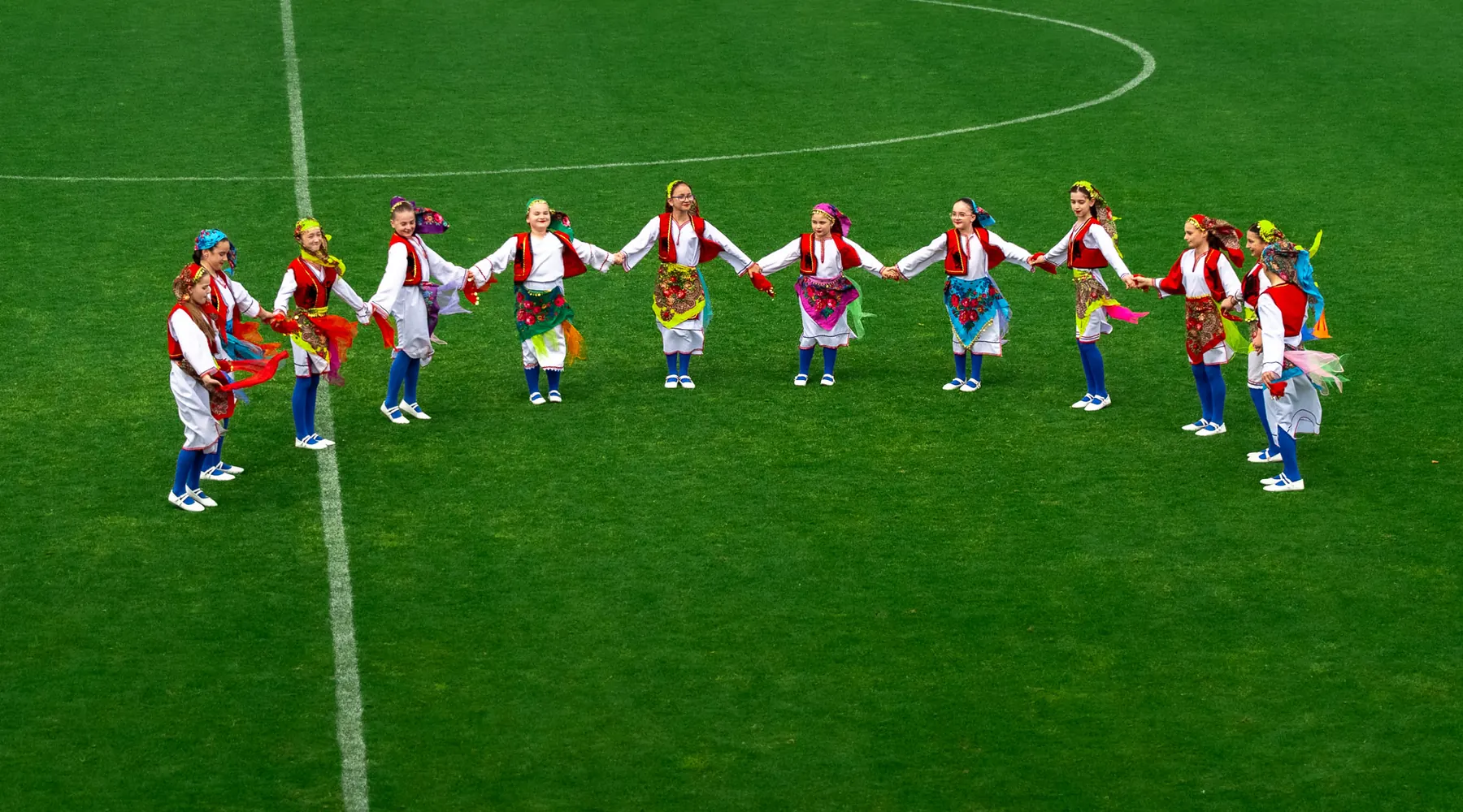A ring of girls in white blouses, red waistcoats and embroidered aprons holds hands on a football pitch