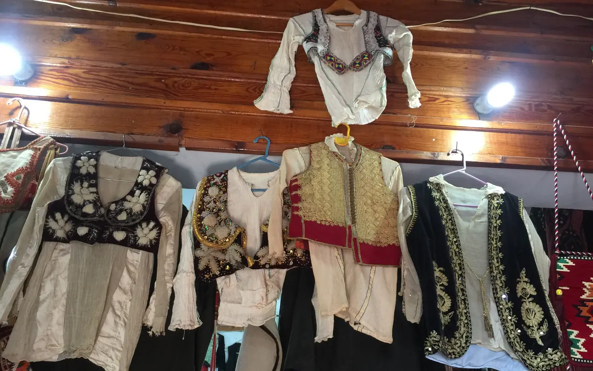 Embroidered blouses and waistcoats in cream, black, red and gold hang on hangers beneath a wooden ceiling