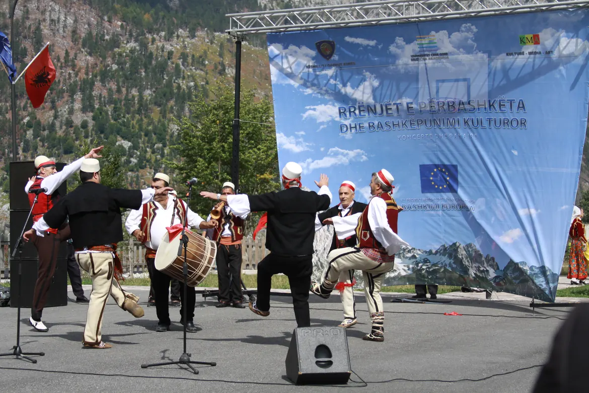 Men in white caps and red-trimmed waistcoats dance beside a large drum and a horn player in front of a stage backdrop