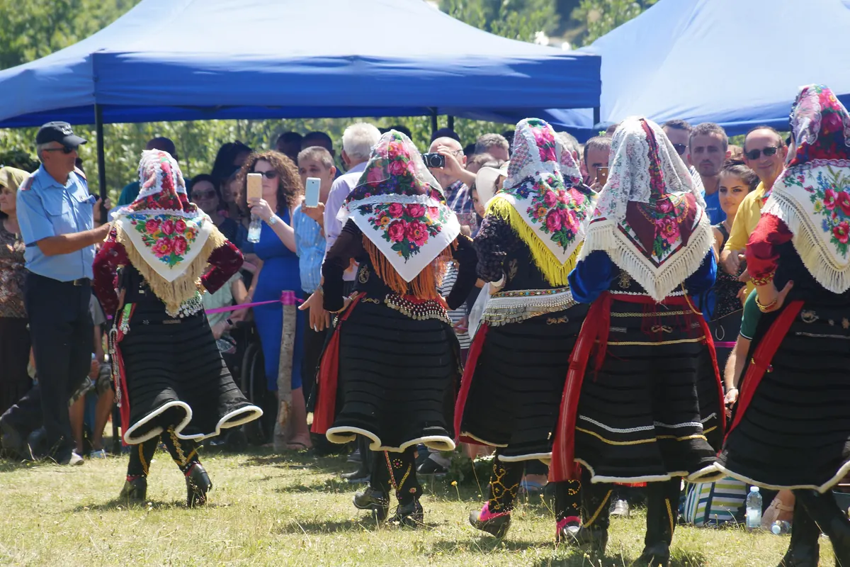 Women in black pleated skirts with red sashes and floral headscarves with gold fringes, seen from behind, dance on grass as a crowd watches