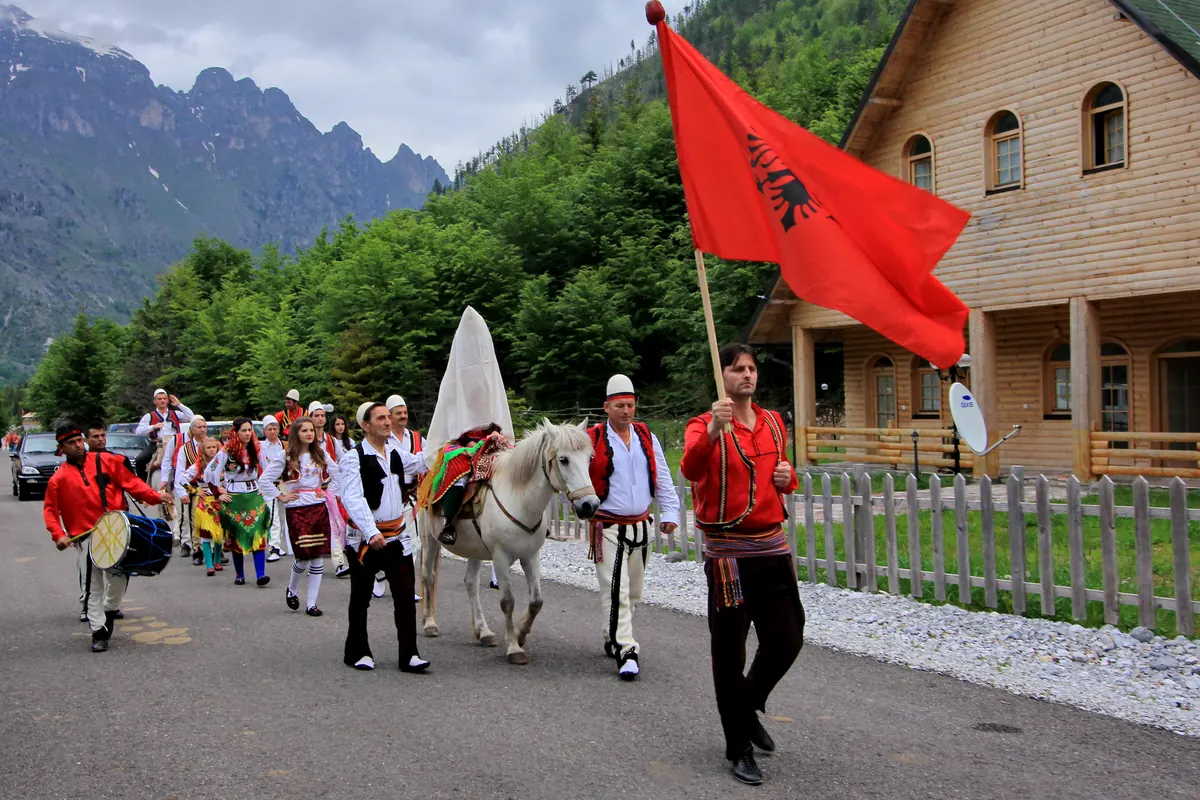 A drummer, a flag bearer and a veiled bride on a white horse lead a procession in folk costume along a road beneath mountains