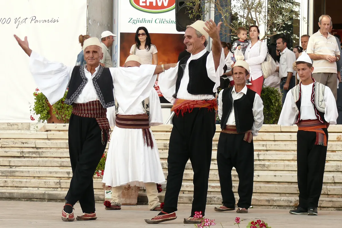 Older men in white shirts, black waistcoats and white felt caps dance with their arms stretched wide in front of a flower-lined stairway