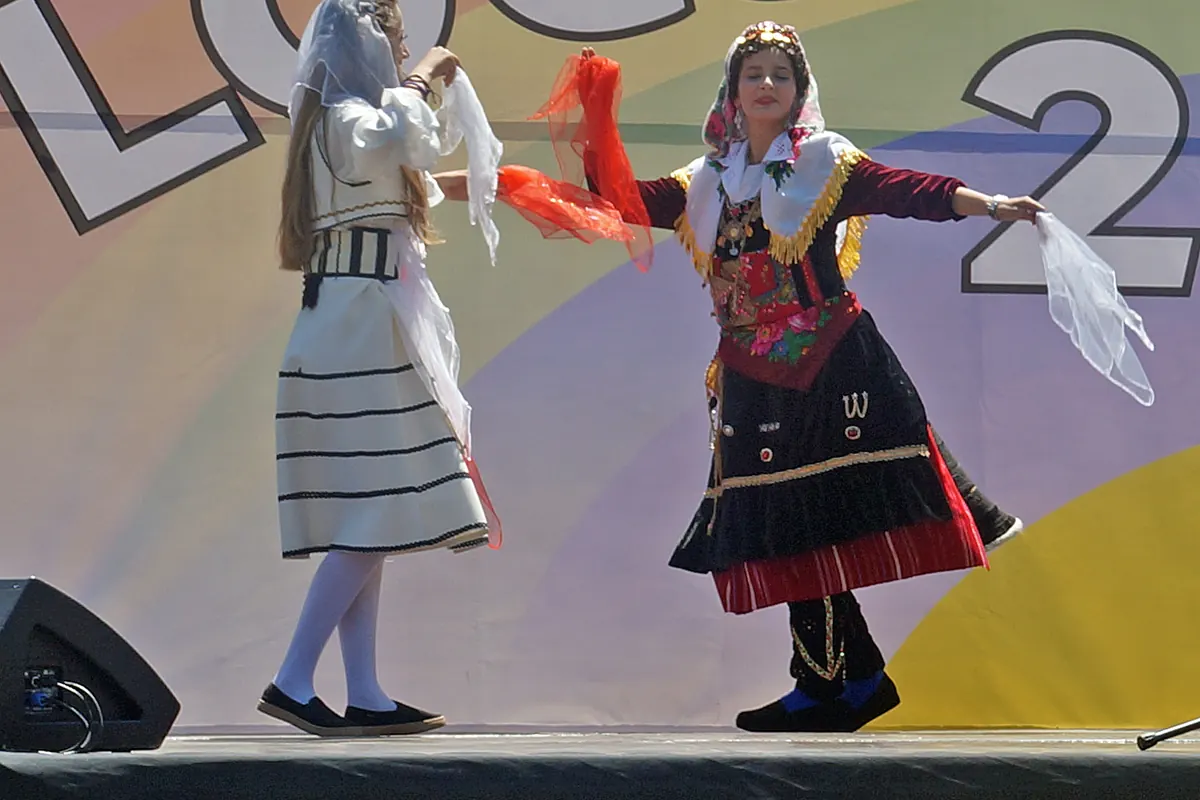 Two dancers in regional costume with long scarves turn towards each other on a stage